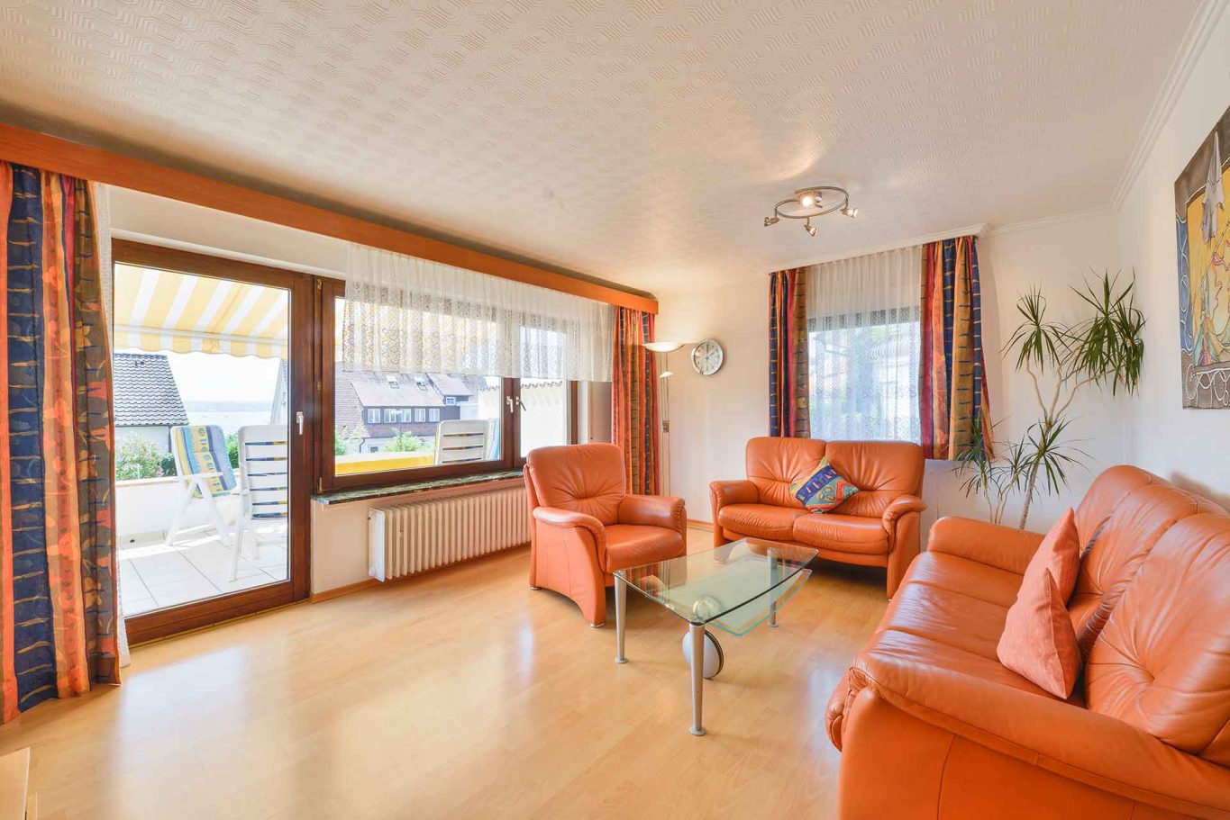 Helles Wohnzimmer mit orangefarbenen Sofas, Glas-Tisch und Fenstern zur Terrasse.