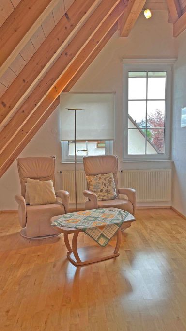 Heller Wohnraum mit zwei Sesseln und einem Tisch unter schrägen Holzdecken.