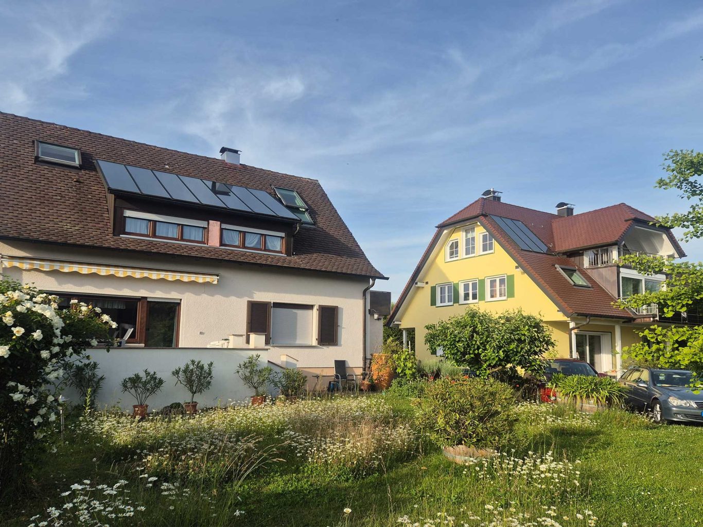 Zwei Häuser mit grünen Gärten, blauer Himmel und Sonnenkollektoren auf dem Dach.
